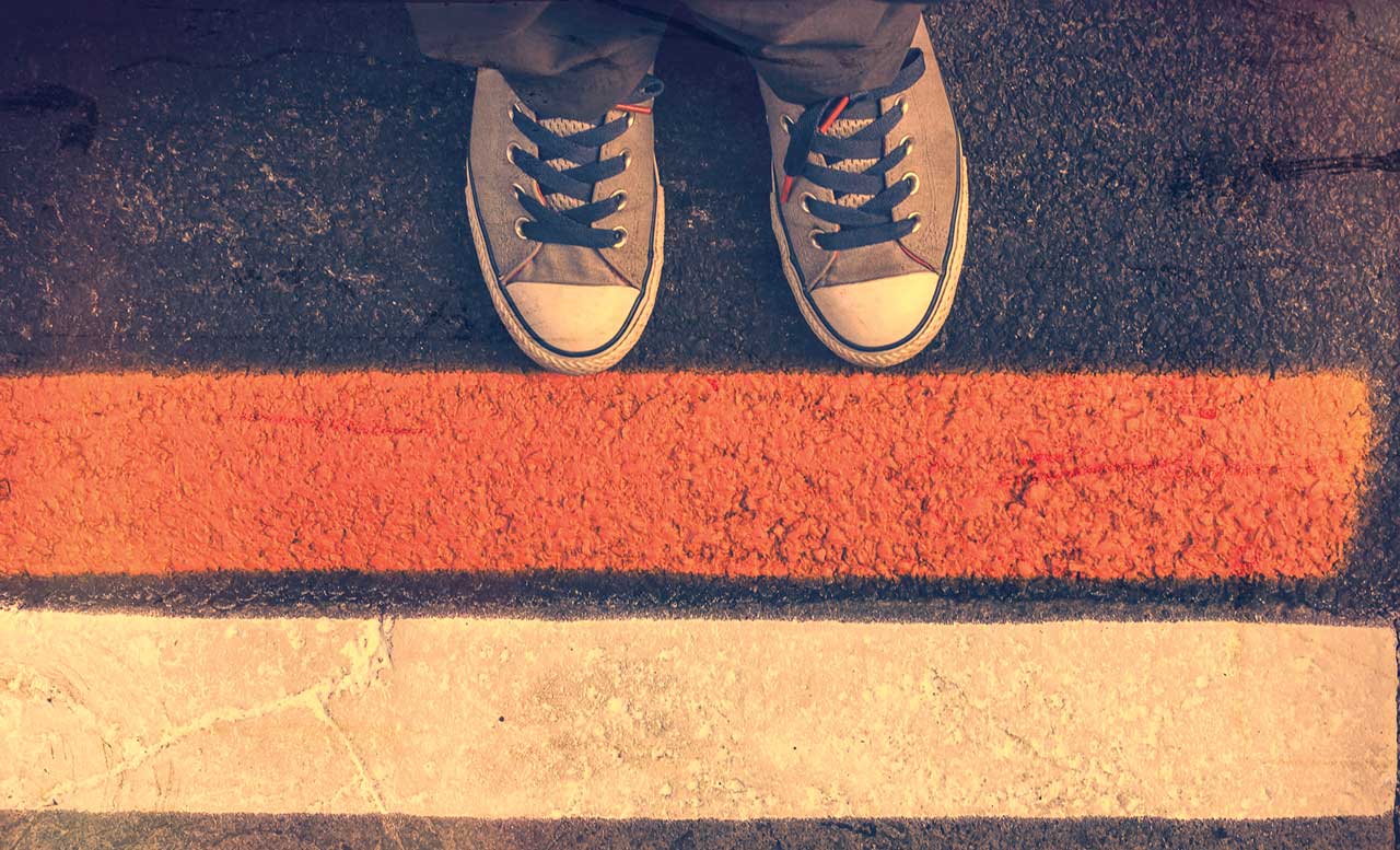 Photo illustration of two lines drawn on asphalt, as seen from above, with the pair of shoes of a person at the edge of the line