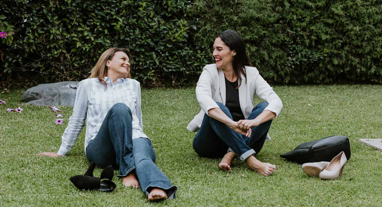 Photo of two women sitting on grass, with their shoes off, taking a break from work