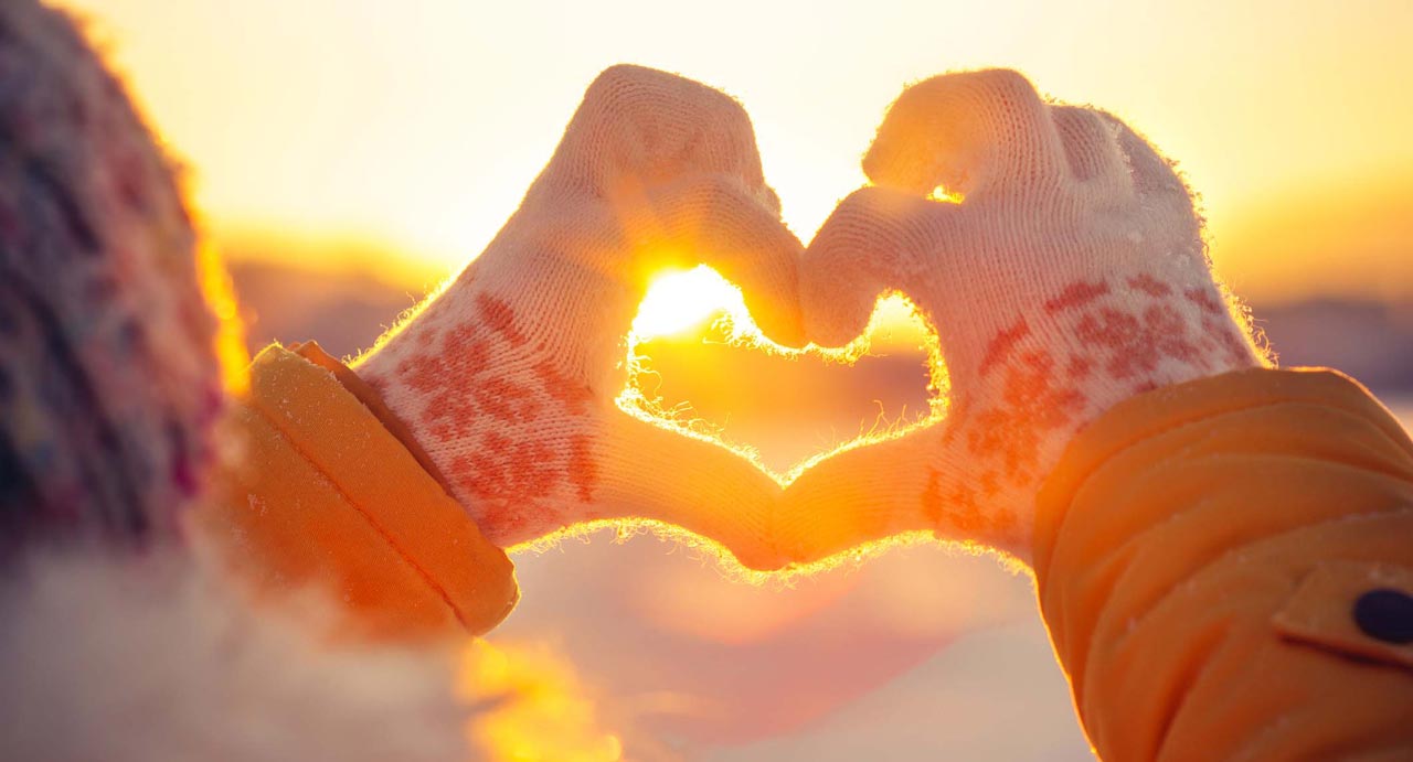 photo closeup of hands wearing winter gloves and forming a heart shape against warm sun rays